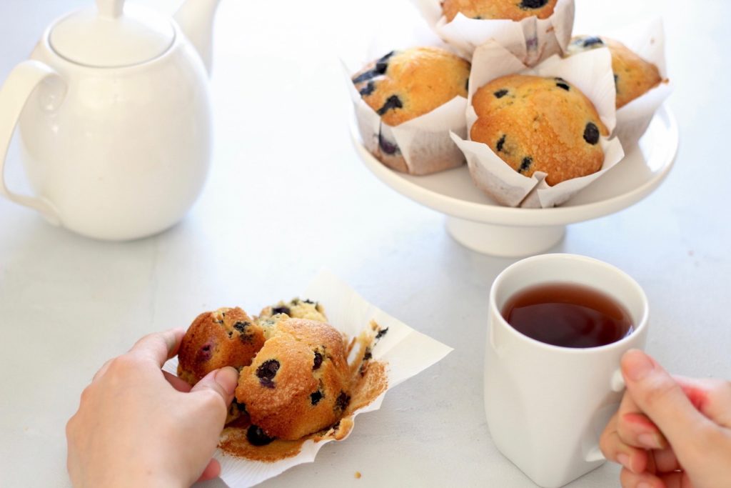 Hands hold a tea cup and a blueberry muffin on a grey table with a small cake stand full of muffins and teapot in the background.