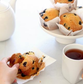 Hands hold a tea cup and a blueberry muffin on a grey table with a small cake stand full of muffins and teapot in the background.