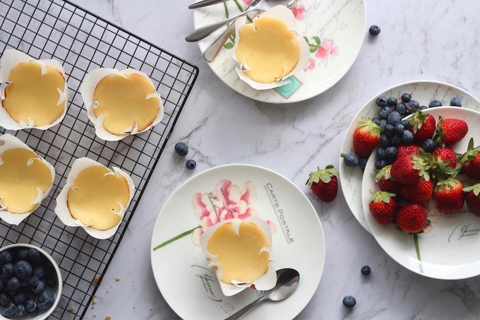 Top-down view of mini baked cheesecakes in white tulip liners on a marble surface, served with fresh strawberries and blueberries.