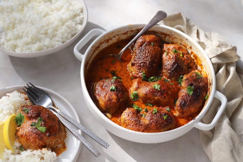 A table setting featuring a white baking dish filled with oven-baked Moroccan chicken thighs. Plates of fluffy white rice, and a serving of the meal surround the main dish.