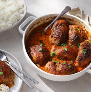 A table setting featuring a white baking dish filled with oven-baked Moroccan chicken thighs. Plates of fluffy white rice, and a serving of the meal surround the main dish.
