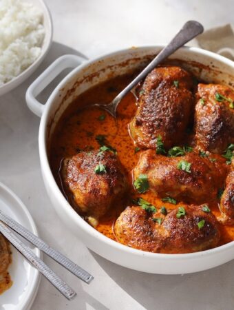 A table setting featuring a white baking dish filled with oven-baked Moroccan chicken thighs. Plates of fluffy white rice, and a serving of the meal surround the main dish.