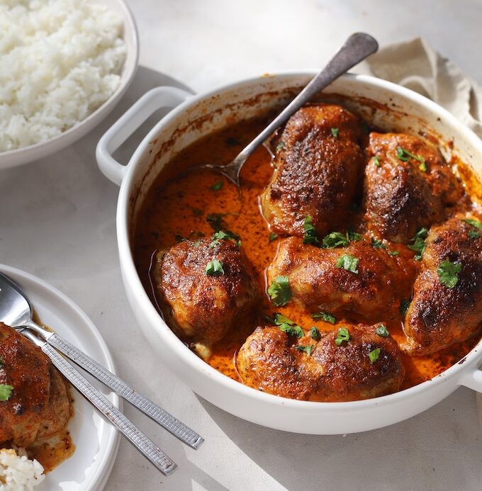 A table setting featuring a white baking dish filled with oven-baked Moroccan chicken thighs. Plates of fluffy white rice, and a serving of the meal surround the main dish.