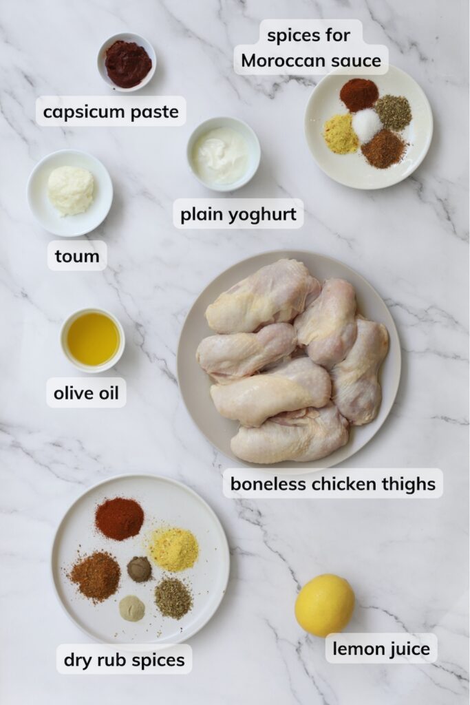 An overhead ingredients shot for oven-baked Moroccan chicken arranged on a marble surface. Labels highlight skin-on chicken thighs, lemon juice, olive oil, capsicum paste, yoghurt, toum, a plate of dry rub spices, and a separate plate of spices for the Moroccan sauce.