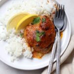 A served plate of oven-baked Moroccan chicken thigh featuring a glossy, caramelised reddish-orange sauce. The chicken is placed alongside a bed of fluffy white rice, garnished with fresh parsley leaves and two lemon wedges, served with a fork and spoon.