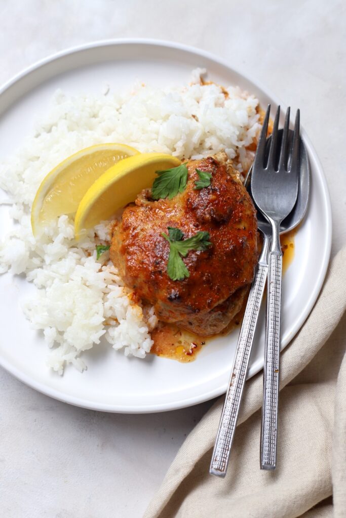 A served plate of oven-baked Moroccan chicken thigh featuring a glossy, caramelised reddish-orange sauce. The chicken is placed alongside a bed of fluffy white rice, garnished with fresh parsley leaves and two lemon wedges, served with a fork and spoon.