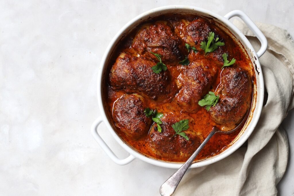An overhead view of finished oven-baked Moroccan chicken thighs in a white ceramic roasting dish. The succulent chicken is coated a Moroccan sauce, garnished with fresh parsley leaves, and served with a metal spoon.