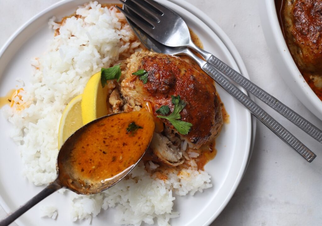 A close-up action shot of a metal spoon drizzling rich Moroccan chicken sauce over an oven-baked chicken thigh. The chicken is served on a white plate with a bed of fluffy white rice, garnished with lemon wedges and fresh parsley.