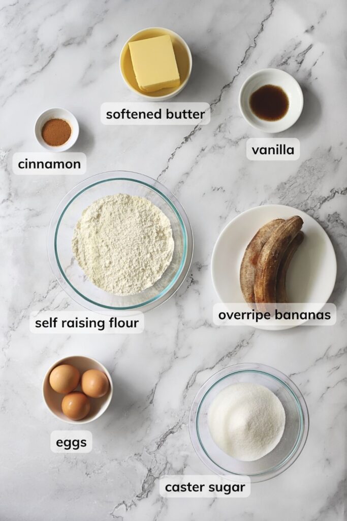 Ingredients for a banana cake recipe arranged on a marble countertop. Labelled ingredients include self raising flour, overripe bananas, caster sugar, eggs, softened butter, vanilla, and cinnamon.