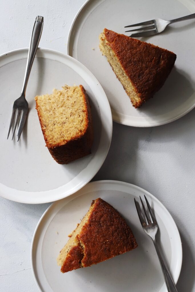 Three slices of homemade one bowl banana cake served on individual plates with forks, showing both the golden-brown crust and the soft, moist interior crumb.