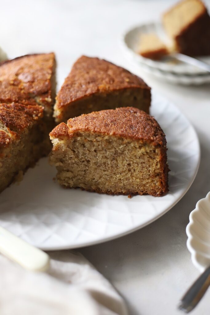 A close-up view of a sliced banana cake on a white textured serving plate, with one thick wedge pulled forward to showcase the soft, moist interior crumb.