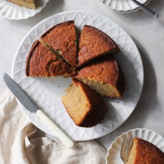 A freshly baked easy banana cake sliced into wedges on a white serving plate. Individual slices of the homemade banana cake are plated on smaller dishes with forks and a dollop of cream.