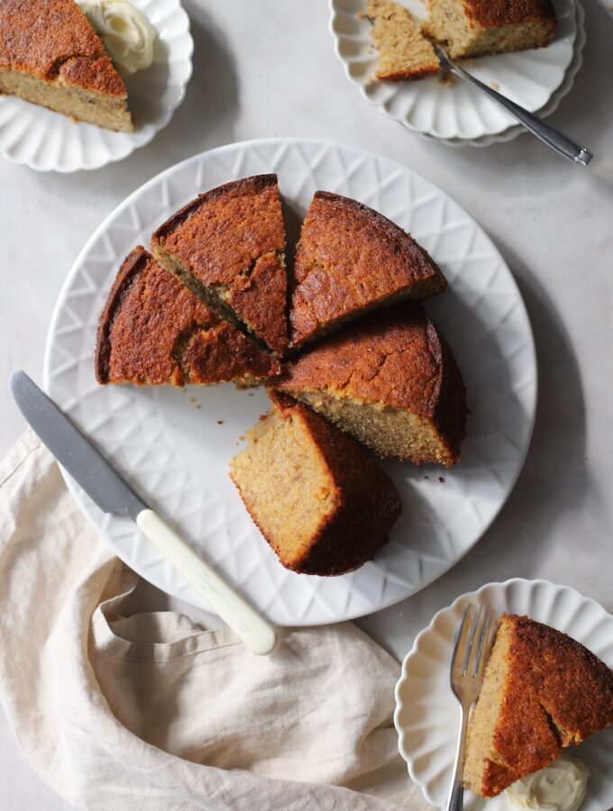 A freshly baked easy banana cake sliced into wedges on a white serving plate. Individual slices of the homemade banana cake are plated on smaller dishes with forks and a dollop of cream.