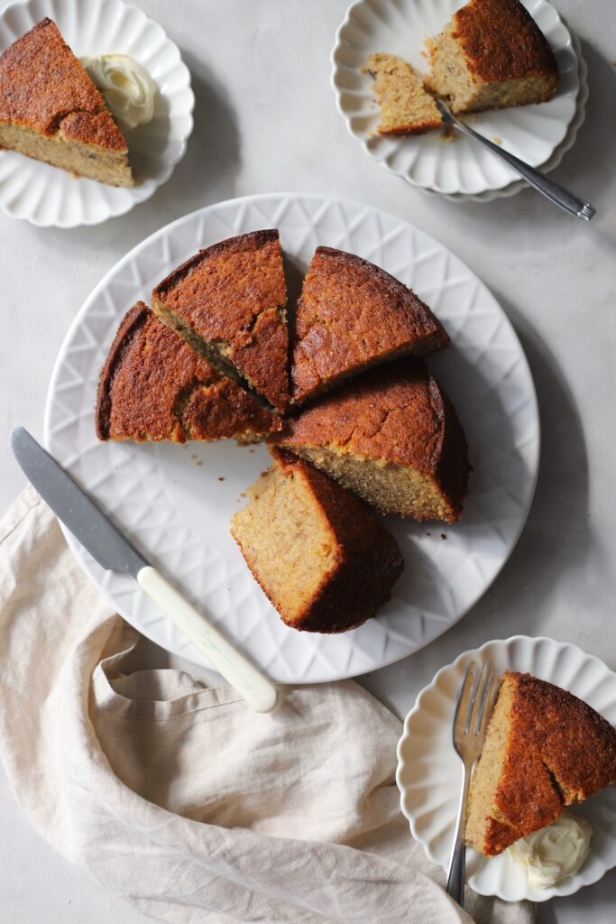 A freshly baked easy banana cake sliced into wedges on a white serving plate. Individual slices of the homemade banana cake are plated on smaller dishes with forks and a dollop of cream.