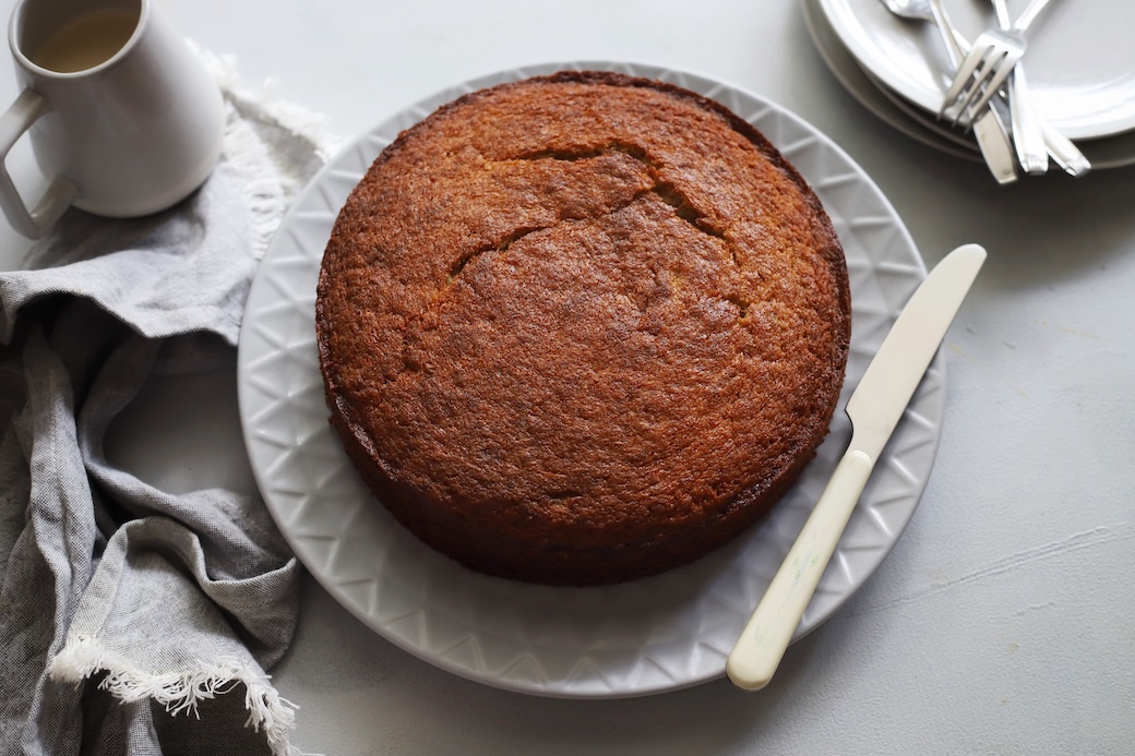 A whole freshly baked moist one bowl banana cake resting on a white serving plate with a knife, ready to be sliced alongside serving plates and a napkin.