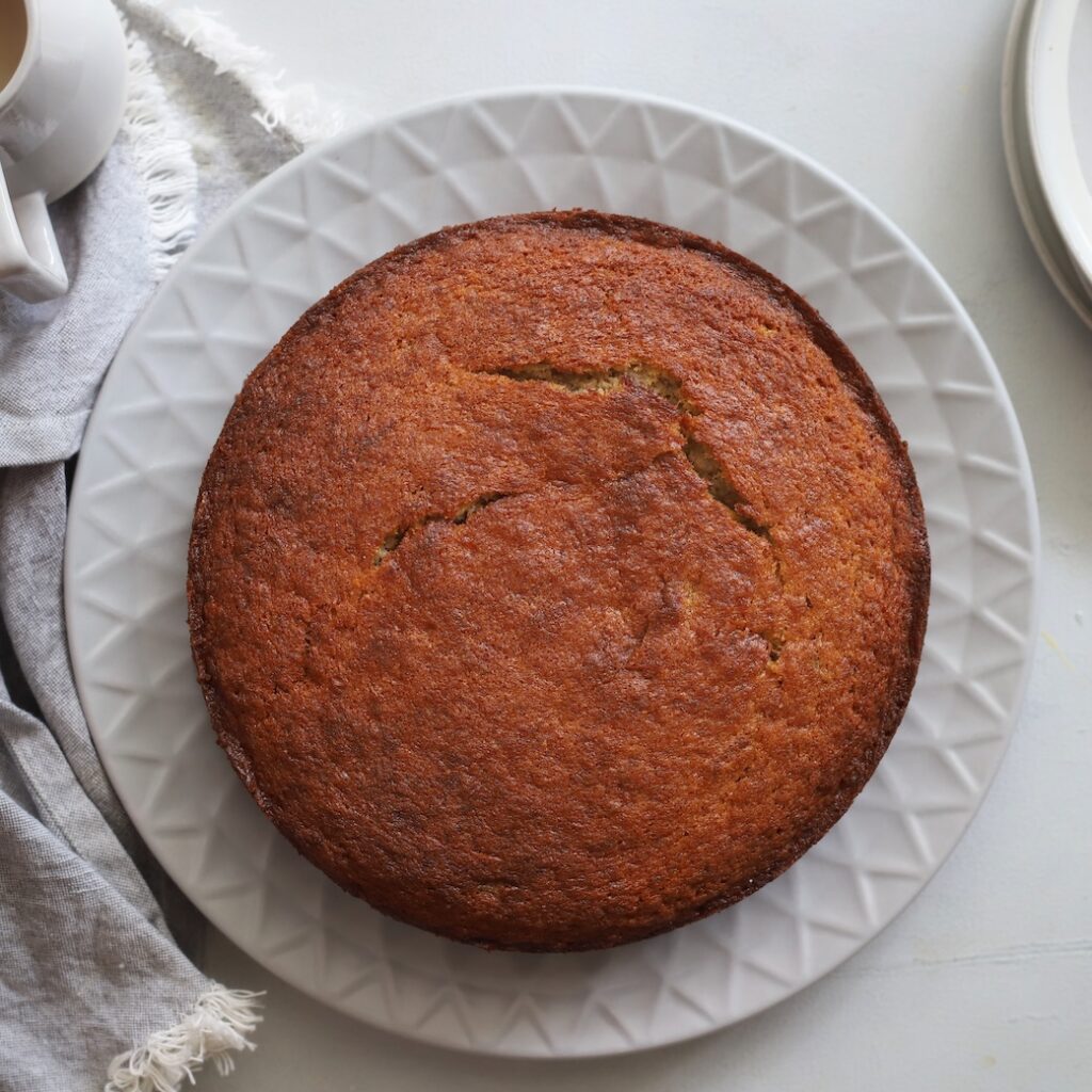 An overhead view of a whole, freshly baked golden-brown banana cake resting on a large white textured serving plate.