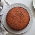 An overhead view of a whole, freshly baked golden-brown banana cake resting on a large white textured serving plate.