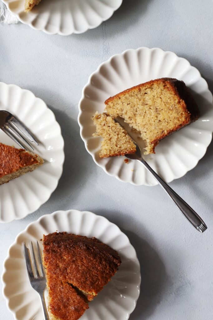 Slices of banana cake served on small white plates with silver forks. The central slice rests on its side, with a fork breaking off a piece to reveal the soft, moist crumb.