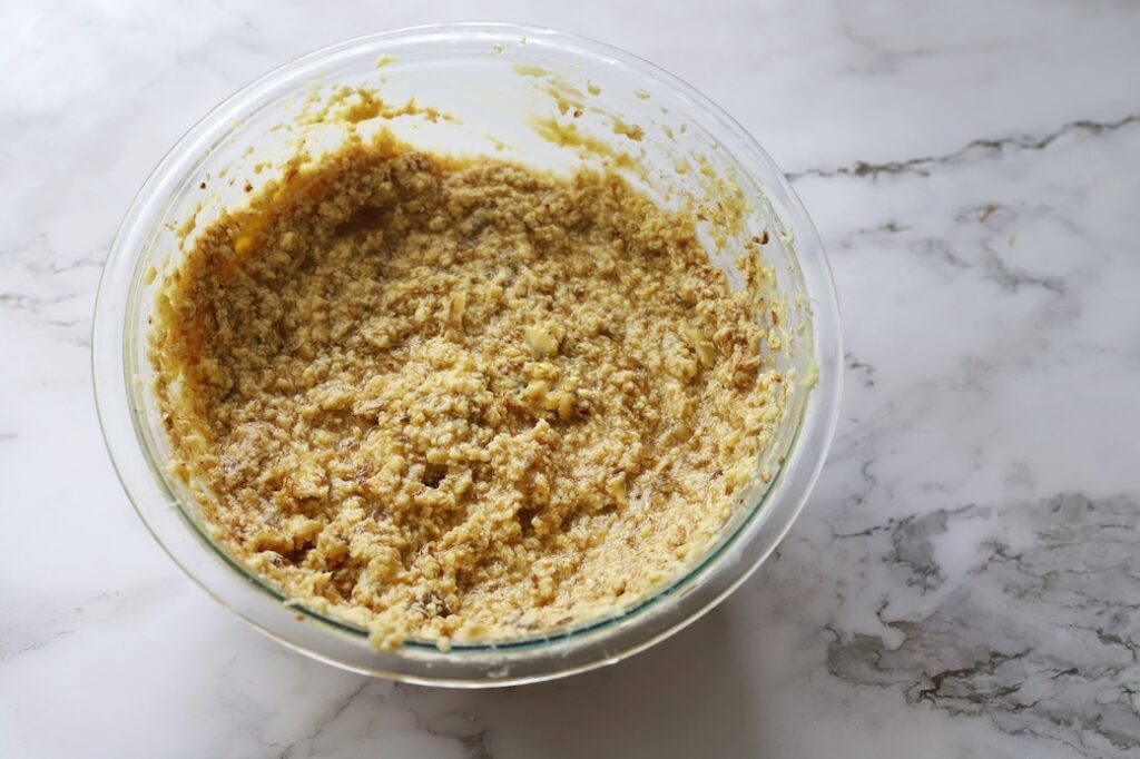A top-down view of a glass mixing bowl containing wet banana cake batter that appears lumpy and temporarily separated, resting on a white marble countertop.