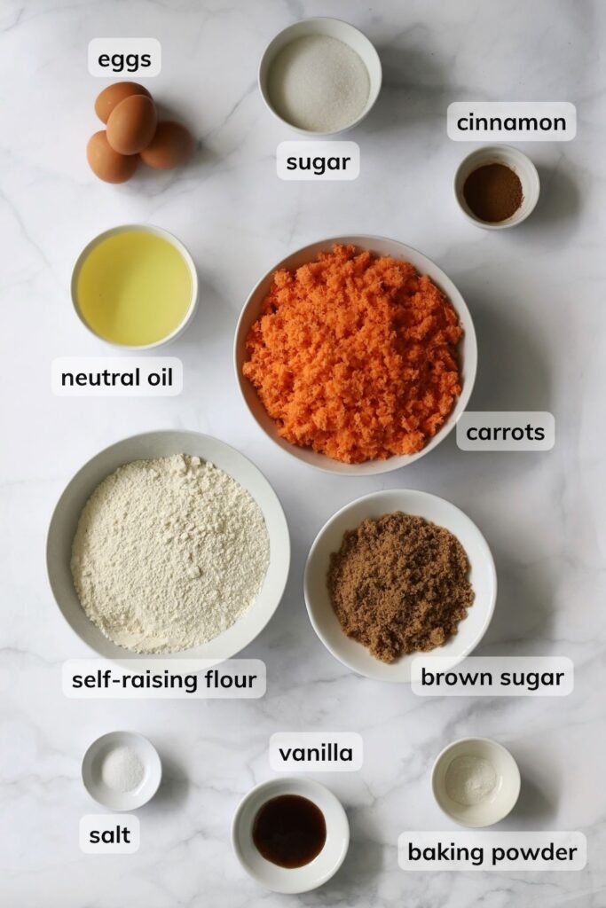 Overhead view of labeled ingredients for a nut-free carrot sheet cake arranged on a marble countertop. Bowls contain finely grated carrots, self-raising flour, neutral oil, brown sugar, white sugar, cinnamon, baking powder, salt, and vanilla, alongside four whole eggs.