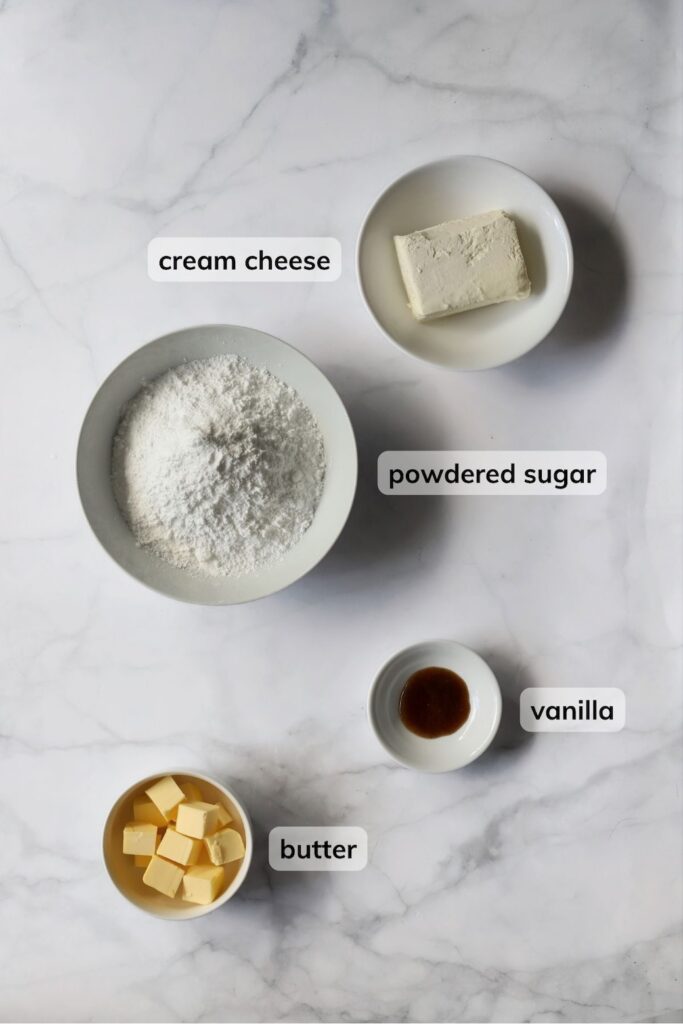 Overhead view of labeled ingredients for a homemade cream cheese frosting arranged on a marble countertop. Four white bowls contain a block of cream cheese, powdered sugar, cubed butter, and vanilla.