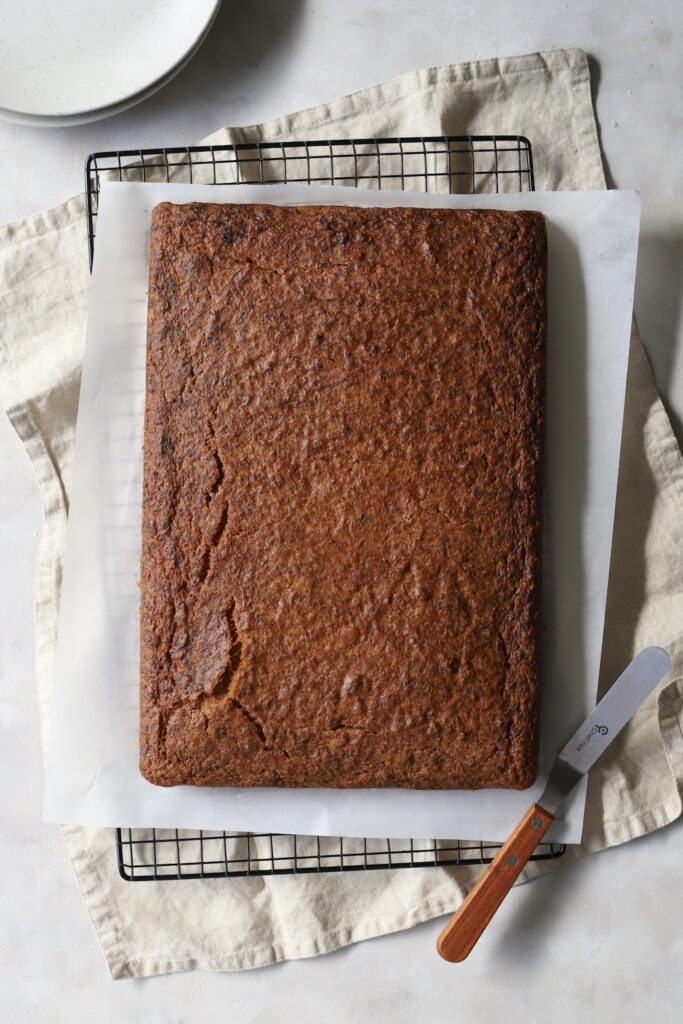 Overhead view of a freshly baked, unfrosted spiced cake resting on parchment paper and a wire cooling rack. A wooden-handled offset spatula sits nearby on a linen cloth, ready for decorating.